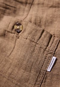 Tela de espiga marrón con un botón y una pequeña etiqueta con el texto "LINDVERGH". Superficie texturizada con costuras visibles y detalle de bolsillo.