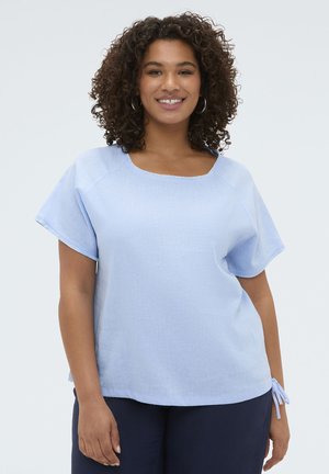 Smilende kvinde med krøllet hår iført en lyseblå kortærmet bluse og navy bukser, stående mod en ensfarvet lys baggrund.