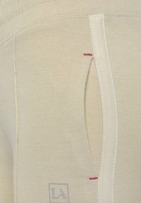 Beige joggerstof met een gladde textuur, voorzien van een zijzakopening, benadrukt door roze stiksels en een klein logo in de rechterbenedenhoek.