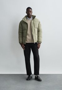 Kurtka puchowa w oliwkowym kolorze z zamkiem z przodu i elastycznymi mankietami, noszona na beżowym swetrze i czarnych spodniach, zestawiona z czarnymi butami.