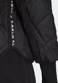 Manche de veste matelassée noire avec poignet côtelé, tissu contrastant en dessous, et texte blanc sur une bande noire près de la zone de la fermeture éclair.