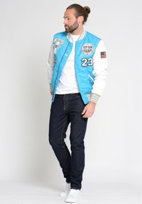 Blauwe bomberjack met witte mouwen, voorzien van patches en geborduurde details. Gepaard met donkere jeans en witte sneakers, staande pose.