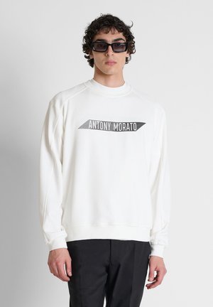 Jonge man die een witte trui draagt met de tekst "Antony Morato" en zwarte broek, staand tegen een effen grijze achtergrond.