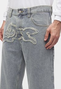 Ανοιχτό γκρι τζιν παντελόνι από denim με ανάγλυφο, υφασμάτινο σχέδιο σκύλου κεντημένο στην μπροστινή άνω περιοχή του μηρού.
