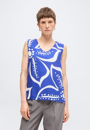 Personne portant un haut bleu sans manches avec de grands motifs floraux blancs et un pantalon gris, debout devant un fond clair uni.