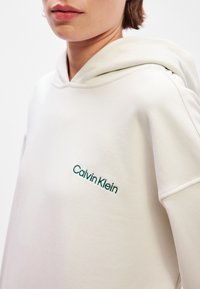 White hooded sweatshirt made of soft fabric, featuring a front pocket and teal "Calvin Klein" embroidery on the left chest.
