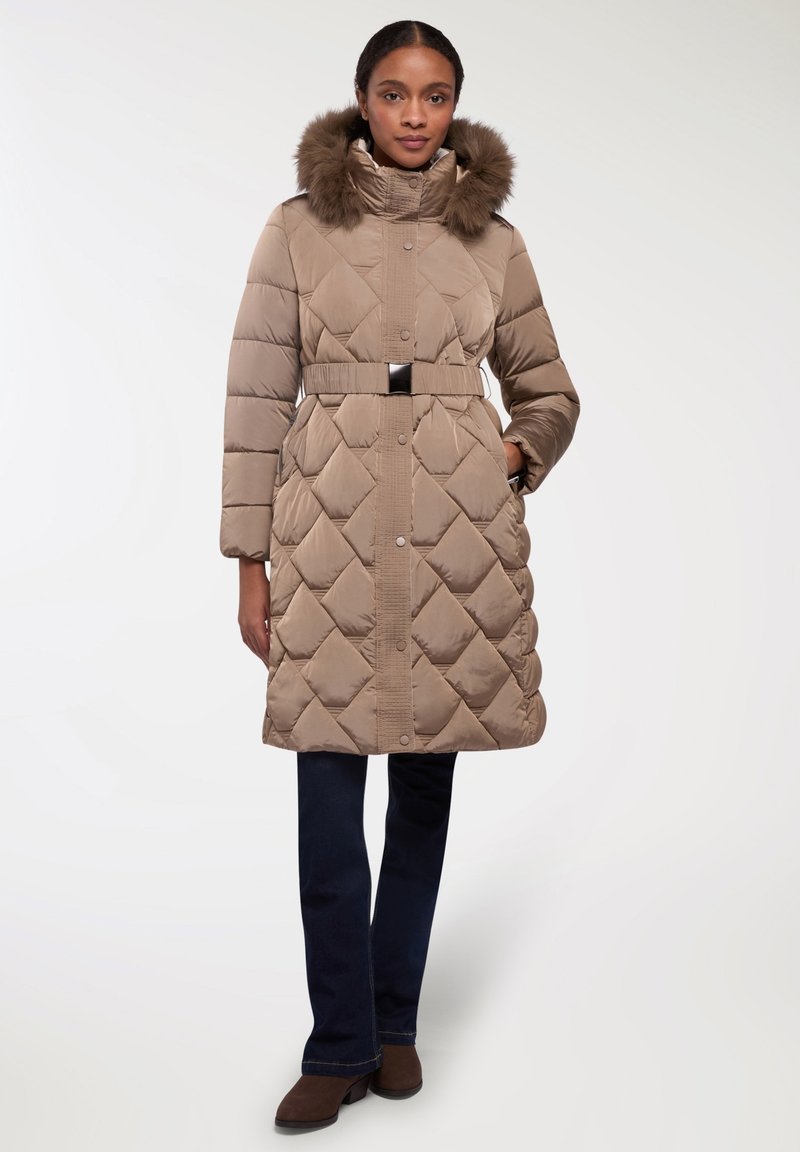 Cappotto imbottito beige con design a rombi, cappuccio con bordo in pelliccia, vita con cintura e chiusura a bottoni. Abbinato a pantaloni blu scuri e stivali marroni.