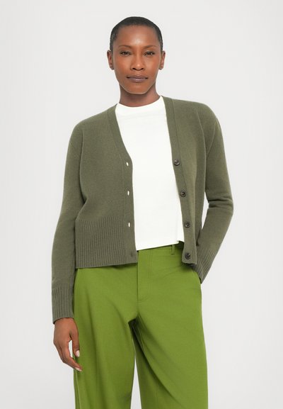 Olive green knitted cardigan with ribbed hem, front buttons, worn over a white shirt. Paired with green trousers, smooth texture.