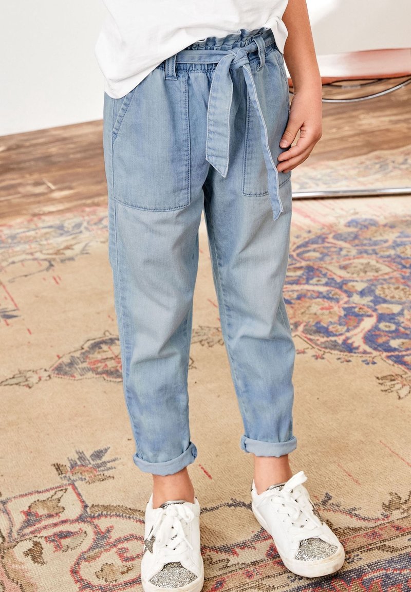 Enfant portant un pantalon en jean bleu clair avec une ceinture nouée à la taille et des revers roulés, associé à des baskets blanches avec des orteils pailletés argentés.