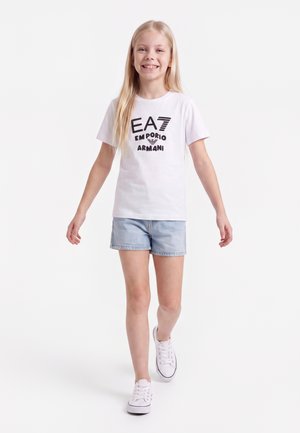 Fille souriante marchant vers l'avant, portant un t-shirt blanc EA7 Emporio Armani, un short en jean bleu clair et des baskets blanches sur fond blanc.