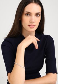 Czarny prążkowany top z krótkimi rękawami; srebrna bransoletka z przezroczystymi kamieniami na nadgarstku; modelka ma długie ciemne włosy i neutralny makijaż.