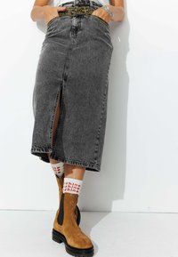 Jupe en denim gris foncé avec une fente avant, ornée de broderies jaunes et noires au niveau de la taille. Une paire de bottines chelsea en daim marron est visible.