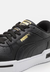 Black leather sneaker with gold accents, featuring a textured surface, lace-up design, and a white rubber sole with branding detail.