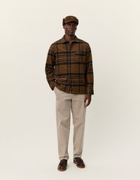 Chemise en flanelle à carreaux marron avec une poche, pantalon en velours côtelé beige et une casquette marron, assortis à des chaussures sombres. Design simple et coupe décontractée.