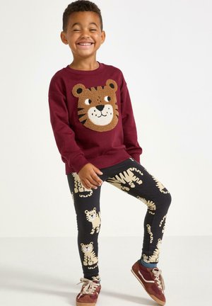 Bordeaux sweatshirt med tigeransigt design, parret med sorte leggings med beige tiger mønstre. Barnet har brune sneakers på.