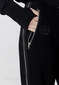 Czarne dresy o smukłym designie, z bocznym zamkiem błyskawicznym, żebrowaną fakturą i okrągłą naszywką z logo. Miękka tkanina i funkcjonalne kieszenie.