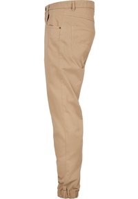Beige tapered-housut, joissa on elastiset hihnat, valmistettu pehmeästä puuvillaseoksesta, sivutaskut ja yksi takatasku napitusyksityiskohdalla.