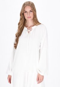Weißes Blusenshirt mit Rüschenausschnitt und Bindedetail. Hat lange, glockenförmige Ärmel und einen glatten, strukturierten Stoff.