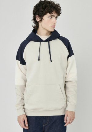 Haut à capuche beige clair avec des accents marine, manches raglan, poche avant, capuche avec cordon de serrage réglable, en tissu doux, design décontracté.