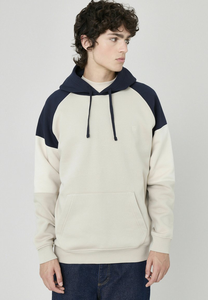 Haut à capuche beige clair avec des accents marine, manches raglan, poche avant, capuche avec cordon de serrage réglable, en tissu doux, design décontracté.