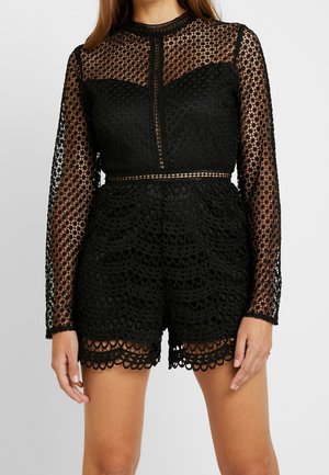 Žena v černém krajkovém overal se sametovými dlouhými průsvitnými rukávy a složitými květinovými vzory, stojící před jednoduchým světlým pozadím.