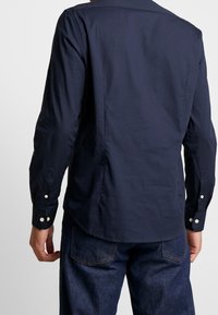 Tummansininen pitkähihaisen paidan, jossa on takayoke ja nappihiukset. Valmistettu sileästä kankaasta, jossa on hienovarainen tekstuuri ja klassinen muotoilu.