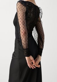 Abito nero con maniche lunghe in chiffon a pois, un corpetto in pizzo e una gonna in satin liscia. Presenta delicate rifiniture in pizzo sui polsini.
