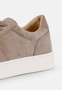 Beige semsket sko med lav profil, rund tå, hvit gummisåle og minimale sømdetaljer. Innvendig har den en myk, tan-farget foring.