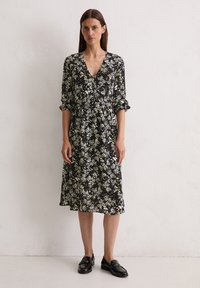 Schwarz gemustertes Kleid mit tiefem V-Ausschnitt, langen Ärmeln mit umgeschlagenen Manschetten und fließender Silhouette. Kleine weiße und gelbe Blumenmuster.