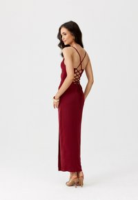 Žena v bezramenných vínově červených šatech s krajkovým detailem na zádech, s zlatými kruhovými náušnicemi, náramky a nahými vysokými podpatky, pózující bokem.