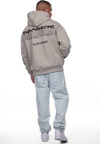 Hellgrauer Hoodie mit einem großen schwarzen Textdesign auf der Rückseite. Kombiniert mit hellblauen Jeans und weißen Sneakern. Lässige, entspannte Passform.