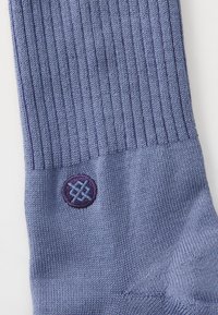 Calza a coste viola con un logo ricamato rotondo in viola scuro e azzurro chiaro sul polsino. Texture liscia con un design strutturato.