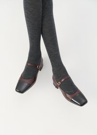 Sapatos Mary Jane em couro borgonha com biqueira em verniz preto, pequeno salto e tira com fecho. Combinados com meias caneladas cinza-escuras.