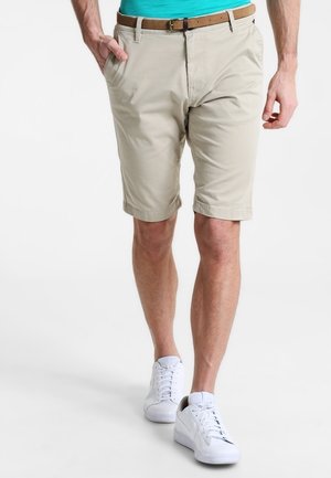 Mężczyzna ubrany w beżowe szorty do kolan z brązowym paskiem, turkusową koszulę i białe tenisówki, stojący z jedną ręką w kieszeni.