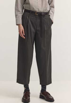Mann trägt hochtaillierte, weit geschnittene, dunkelgraue plissierte Hose mit schwarzem Gürtel, braune strukturierte Lederschuhe und gemusterte Socken.
