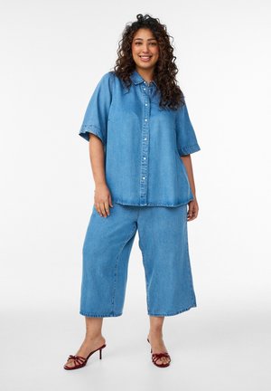 Kvinde med krøllet hår, smilende, iført en løs denim skjorte med knapper, vide, cropped denimbukser og vinrøde hælede sandaler, stående på hvid baggrund.