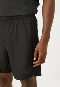 Artimiausias kadras asmens, vilkintis juodais sportiniais šortais su elastiniu juosmeniu, ant kairės kojos apačios matomas EA7 Emporio Armani logotipas.
