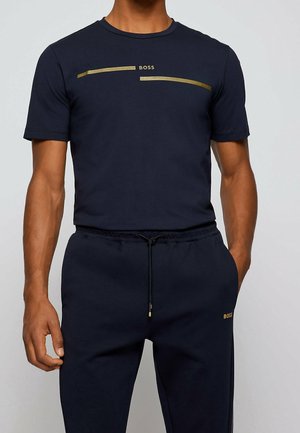 Homme portant un t-shirt bleu marine foncé et un pantalon de survêtement assorti avec le logo "BOSS" doré sur la poitrine et la cuisse. Les mains sont détendues le long du corps.