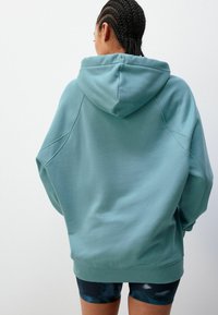 Haut à capuche surdimensionné, vert menthe en tissu doux avec cordon de serrage, épaules tombantes et poignets côtelés. Porté par-dessus un short noir à motifs.