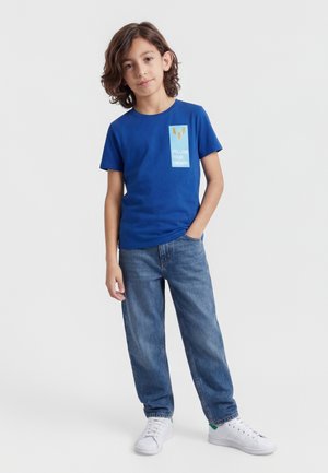 Un garçon aux cheveux mi-longs se tient debout, portant un T-shirt bleu "Suis tes rêves", un jean bleu et des baskets blanches, sur un fond uni.