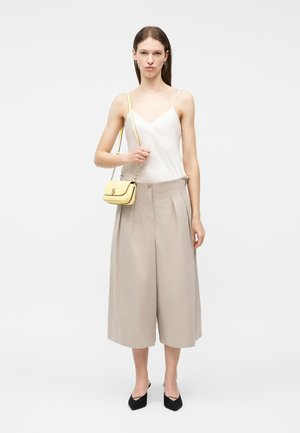 Mujer con top sin mangas blanco, pantalones beige de pierna ancha y corte cropped, tacones negros, y sosteniendo un pequeño bolso de hombro amarillo con cadena dorada.