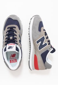 Un paio di sneakers New Balance 574 in grigio, blu navy e rosso, con tomaia in mesh e suede, viste dall'alto e di lato su uno sfondo bianco.