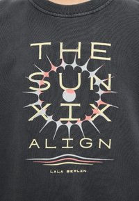 Μαύρο πουκάμισο με στυλιζαρισμένη ηλιακή ακτινοβολία γύρω από τις λέξεις «THE SUN SIX ALIGN» και το κείμενο «LALA BERLIN» από κάτω.