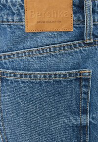 Jeans de mezclilla azul con una etiqueta de cuero marrón que dice "Colección DENIM de Bershka", con costuras estándar en los bolsillos traseros y un ajuste clásico.