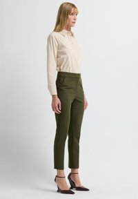 Beige Langarmhemd mit Knöpfen und Kragen, kombiniert mit hoch taillierten olivgrünen Anzughosen und weinroten Absatzschuhen.