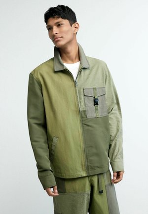 Noor mees, kes kannab oliivirohelist lapitehnikaga jopet ja sobivaid pükse, seisab ja vaatab veidi küljele ühetaustalise tausta eest.