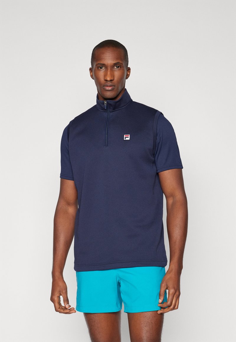 Marineblå kortermet polo med glidelås-krage, med teksturert stoff og liten logo-applikasjon. Sammen med tealfargede shorts.