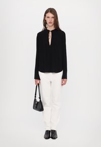 Black long-sleeve blouse with a V neckline and tie detail, featuring embroidered cuffs, paired with white jeans and black studded boots.