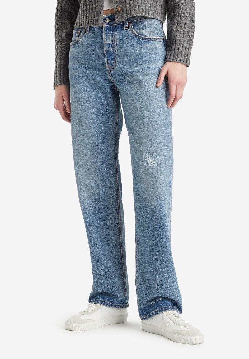 Personne portant un jean droit bleu clair, des baskets blanches et un pull gris à torsades, debout sur un fond blanc.