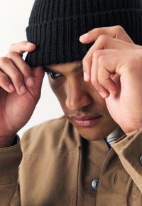 Crna rebrasta pletena kapa koju nosi osoba dok je podešava rukama. Osoba je odjevena u smeđu jaknu, prikazana je izbliza.